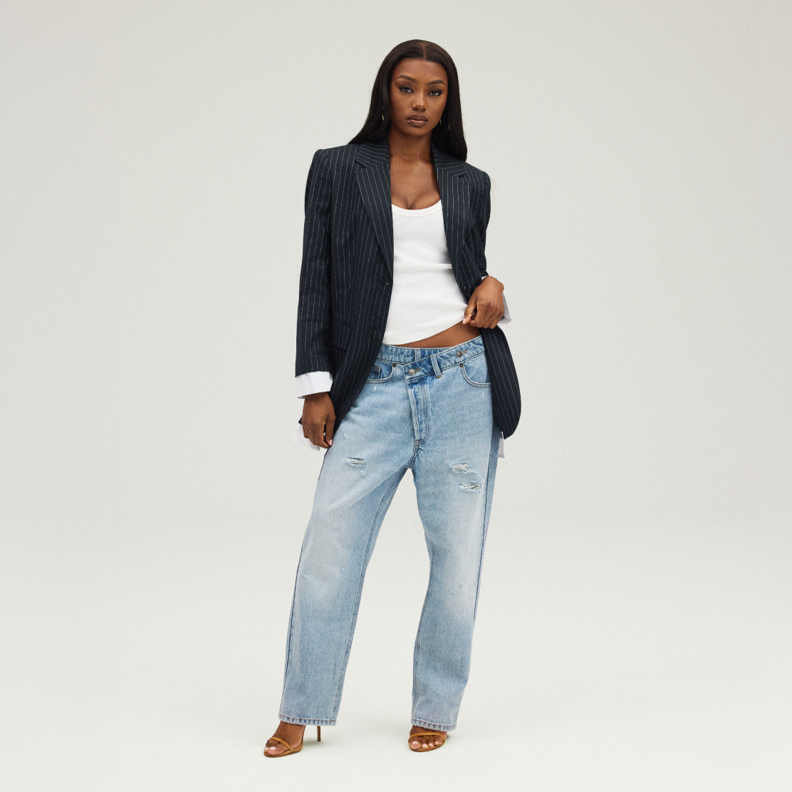 Model wearing a pinstripe blazer with cuffed sleeves over a white ribbed tank top, styled with light-wash, high-rise distressed boyfriend jeans and strappy heeled sandals.