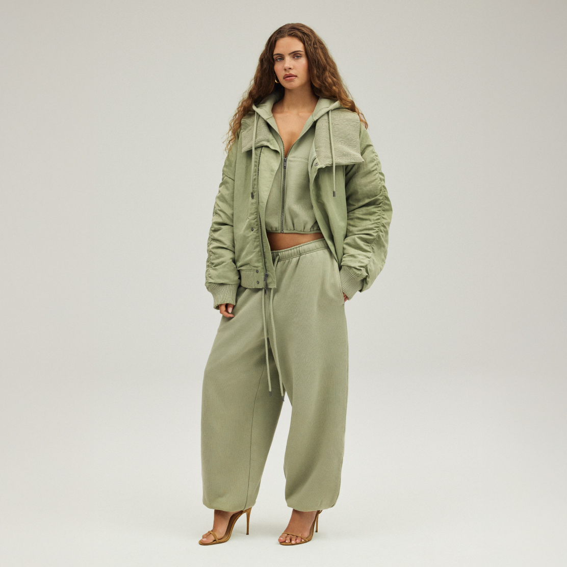 Model wearing an oversized sage green bomber jacket layered over a cropped hoodie and wide-leg sweatpants, styled with strappy heels for a luxe athleisure look.