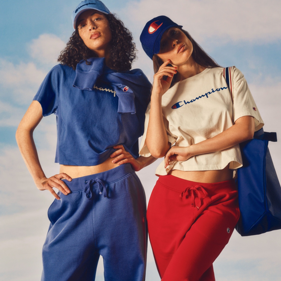 Two people wearing Champion apparel pose against a sky backdrop, one in a blue cropped tee, joggers and cap, the other in a cream tee, red joggers, navy cap and tote