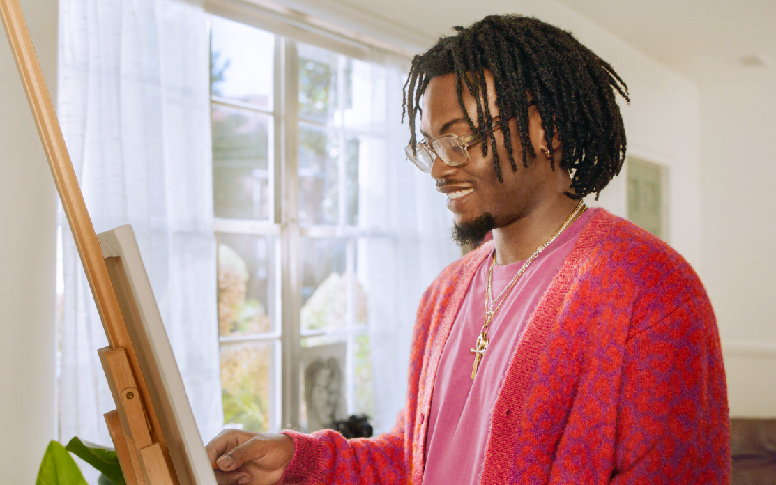A man in a multi-tonal pink sweater stands smiling in front of an easel.