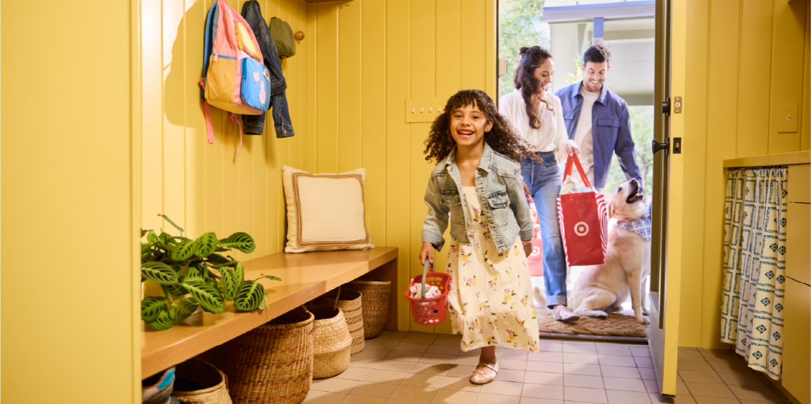 A family walks through the door of their home; a child laughs while carrying a toy Target basket and the parents carry a reusable Target shopping bag.